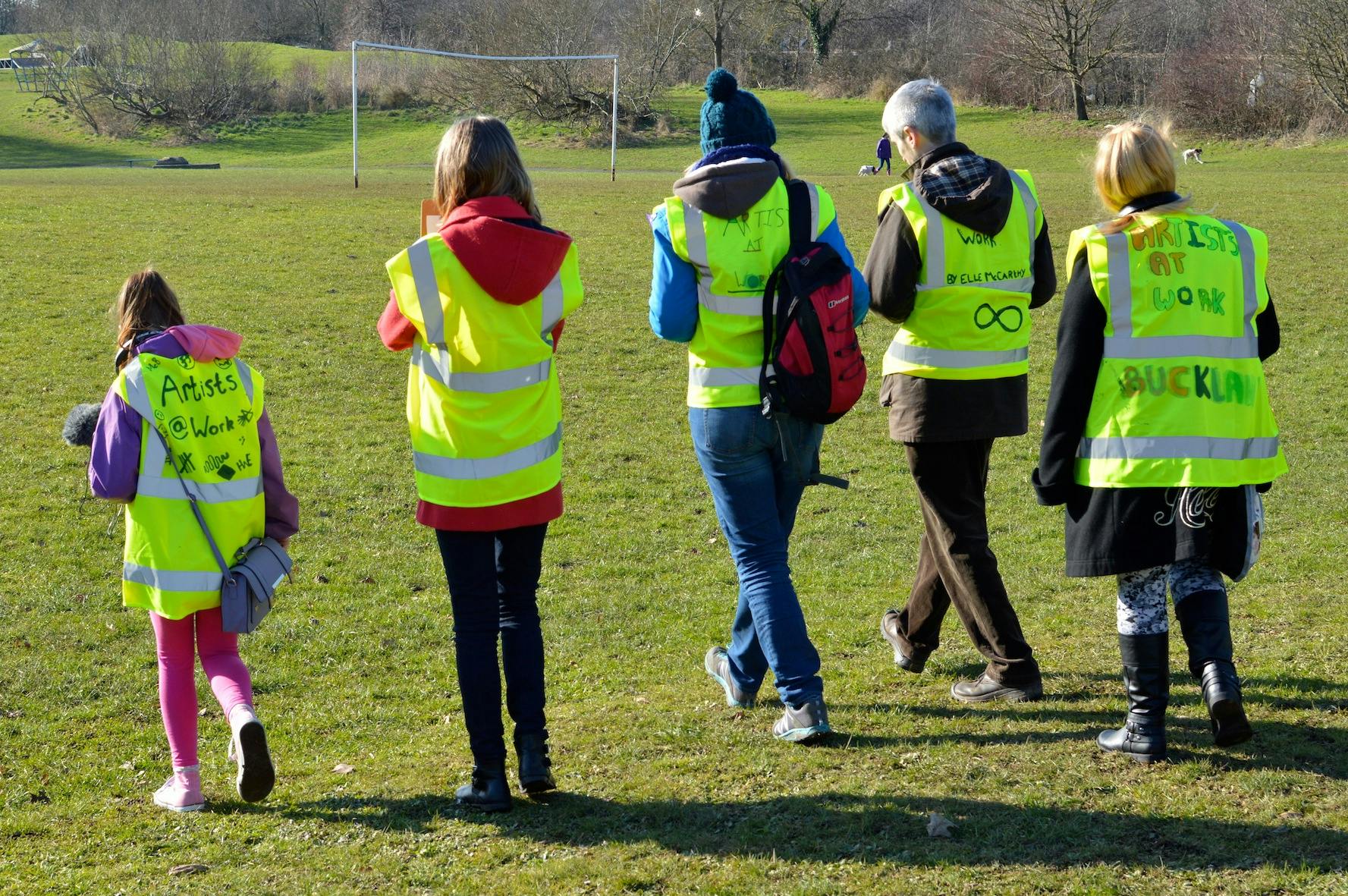 A group of adults and children wearing jackets saying 'artist at work' walking across a playing field